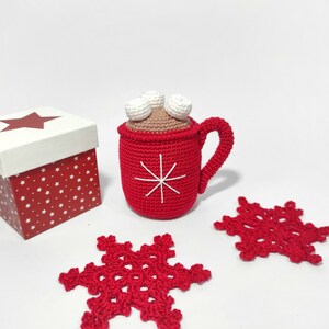 May include: A red crocheted mug with a white snowflake design and white marshmallows on top. The mug is sitting on a white surface next to a red and white gift box and two red crocheted snowflakes.