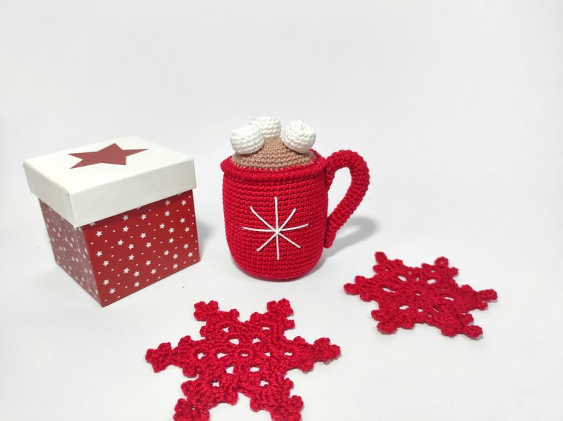 May include: A red crocheted mug with a white snowflake design and white marshmallows on top. The mug is sitting on a white surface next to a red and white gift box and two red crocheted snowflakes.
