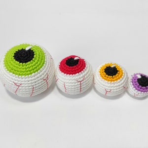 May include: Four crocheted eyeball ornaments in different colors: green, red, yellow, and purple. The eyeballs are all white with black pupils and are arranged in a line from largest to smallest.
