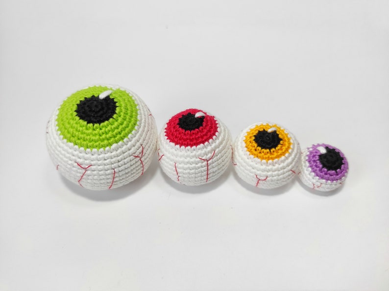 May include: Four crocheted eyeball ornaments in different colors: green, red, yellow, and purple. The eyeballs are all white with black pupils and are arranged in a line from largest to smallest.