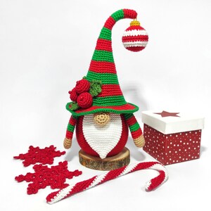 May include: A handmade Christmas gnome with a red and green striped hat and body, a white beard, and a decorative ornament. The gnome is displayed with red snowflakes, a candy cane, and a gift box. The gnome is approximately 10 inches tall.