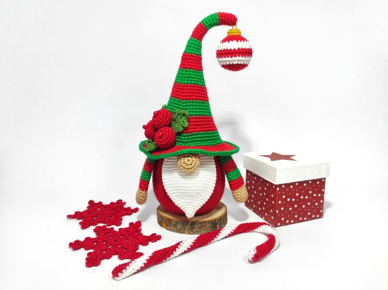 May include: A handmade Christmas gnome with a red and green striped hat and body, a white beard, and a decorative ornament. The gnome is displayed with red snowflakes, a candy cane, and a gift box. The gnome is approximately 10 inches tall.