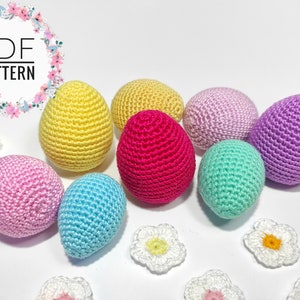 Puede incluir: Un conjunto de ocho huevos de Pascua tejidos a crochet en varios colores, incluyendo amarillo, rosa, azul, verde y morado. Los huevos est&aacute;n dispuestos sobre una superficie blanca con peque&ntilde;as flores tejidas a crochet.