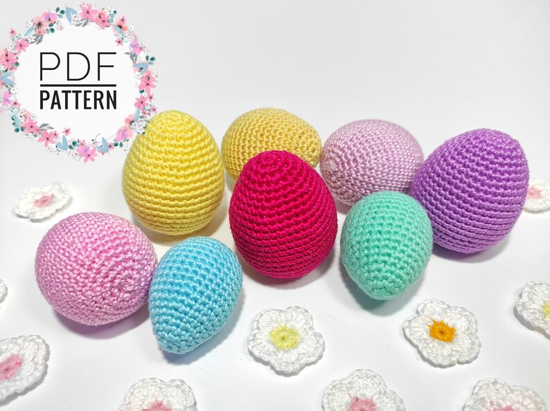 Puede incluir: Un conjunto de ocho huevos de Pascua tejidos a crochet en varios colores, incluyendo amarillo, rosa, azul, verde y morado. Los huevos est&aacute;n dispuestos sobre una superficie blanca con peque&ntilde;as flores tejidas a crochet.