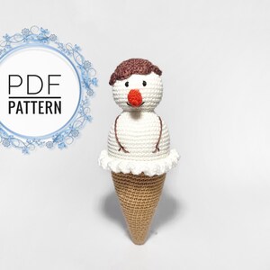 May include: Crocheted snowman ice cream cone amigurumi toy pattern. The snowman has a brown hair, white body, and a red nose. The ice cream cone is brown and white.