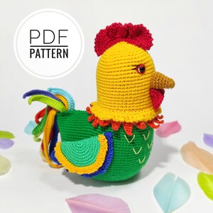 Puede incluir: Un juguete de gallo de crochet con cabeza amarilla, cresta roja, cuerpo verde y cola arcoíris. El juguete está sentado sobre una superficie blanca con plumas de colores esparcidas a su alrededor. El texto "PDF PATTERN" es visible en un círculo en la esquina superior izquierda de la imagen.