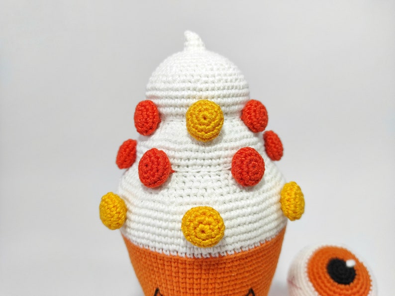 May include: A white crocheted cone-shaped toy with orange and yellow polka dots. The toy has a small orange eye on the side.