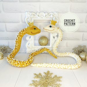 May include: Two crocheted snakes wearing crowns, one is gold and the other is white. The snakes are facing each other and are on a white surface. The text "CROCHET PATTERN" is in a circle in the upper right corner of the image.