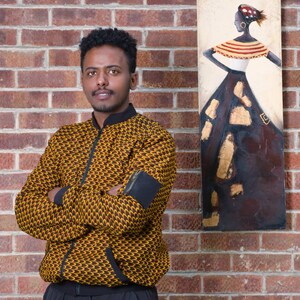 May include: A person wearing a yellow and brown patterned jacket with black sleeves and black pants. The person is standing in front of a brick wall with a painting of a woman in a colorful dress.