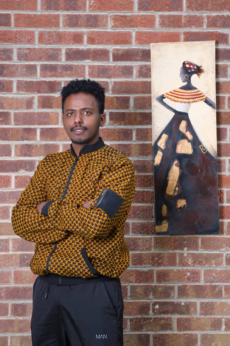 May include: A person wearing a yellow and brown patterned jacket with black sleeves and black pants. The person is standing in front of a brick wall with a painting of a woman in a colorful dress.