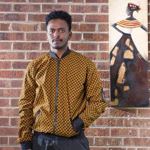 May include: A man wearing a brown and yellow patterned bomber jacket with black pants. He is standing in front of a brick wall with a painting of a woman in a colorful dress on the wall.