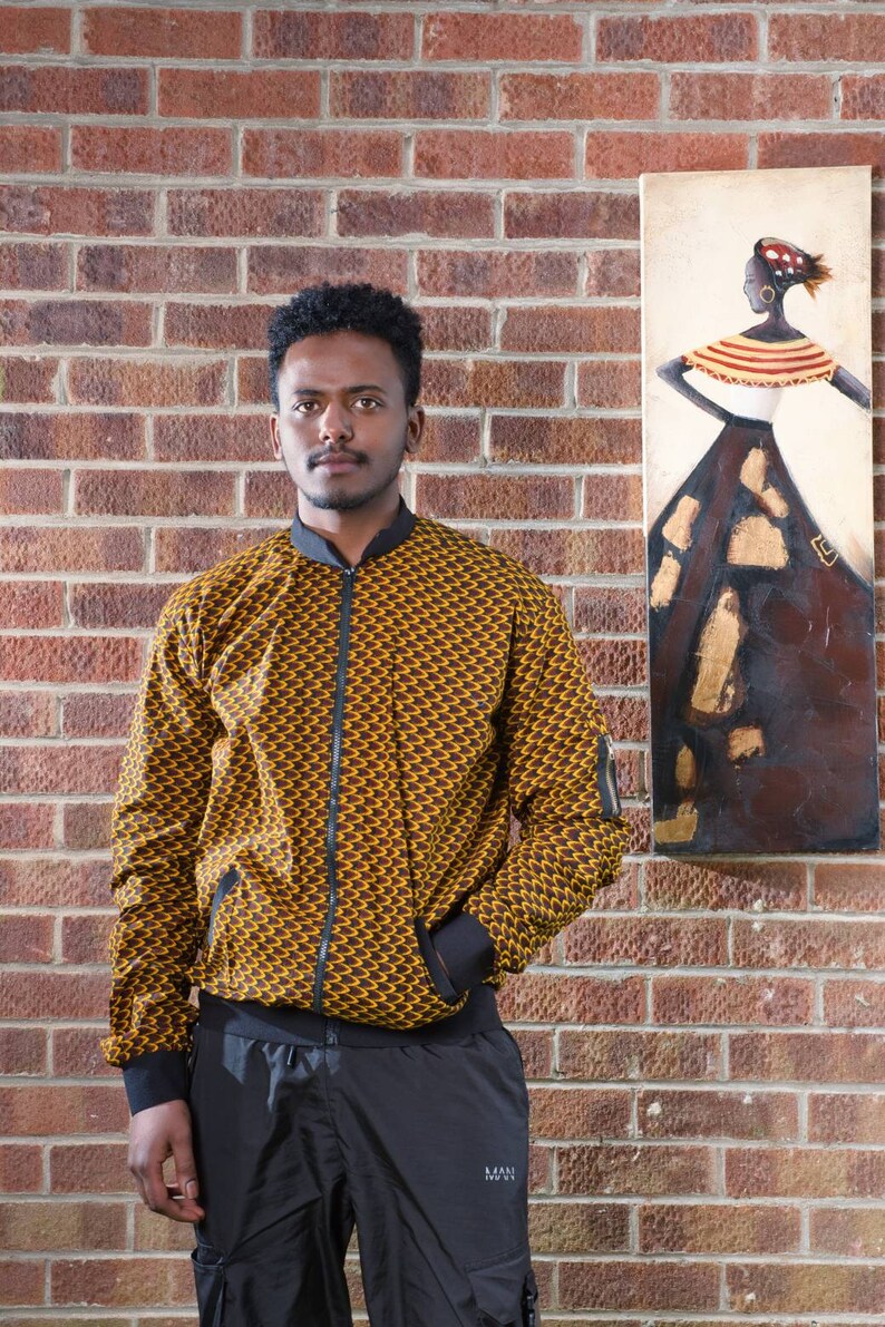 May include: A man wearing a brown and yellow patterned bomber jacket with black pants. He is standing in front of a brick wall with a painting of a woman in a colorful dress on the wall.
