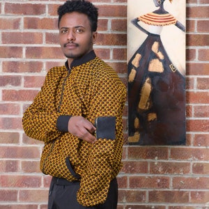 May include: A man wearing a yellow and brown patterned bomber jacket with a black patch on the sleeve. He is standing in front of a brick wall with a painting of a woman in a brown dress.