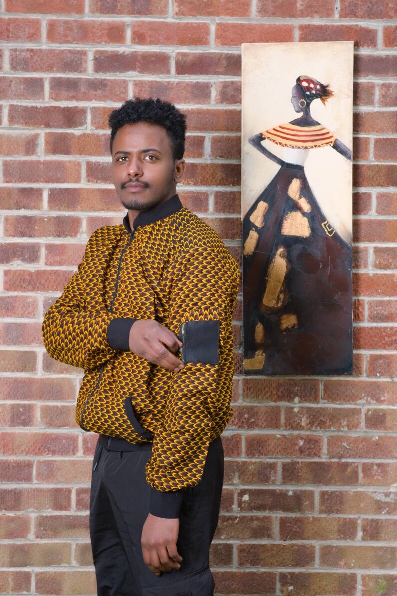 May include: A man wearing a yellow and brown patterned bomber jacket with a black patch on the sleeve. He is standing in front of a brick wall with a painting of a woman in a brown dress.