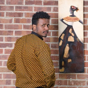 May include: A man wearing a yellow and brown patterned shirt with black pants stands in front of a brick wall. A painting of a woman in a brown dress with gold accents hangs on the wall behind him.