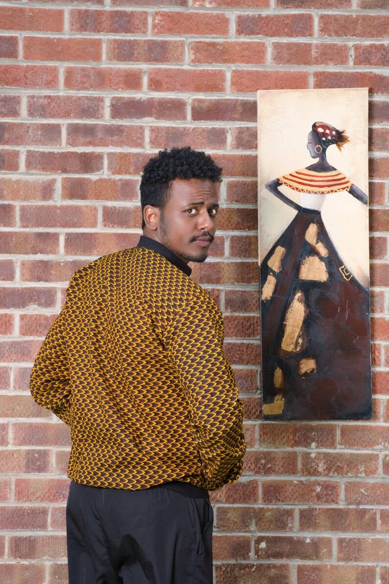 May include: A man wearing a yellow and brown patterned shirt with black pants stands in front of a brick wall. A painting of a woman in a brown dress with gold accents hangs on the wall behind him.
