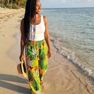 May include: A person wearing a white tank top and colorful patchwork pants walks on a sandy beach. The pants feature a vibrant mix of green, yellow, and orange patterns. A straw hat is held in the left hand.