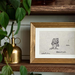 May include: A rectangular wooden picture frame containing a light-colored paper print. The print features a black ink line drawing of a figure emerging from stylized waves, with additional wavy line patterns positioned below the central figure.