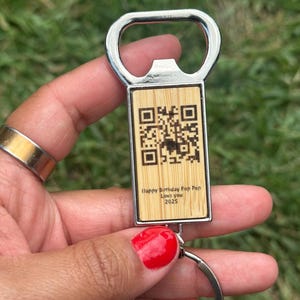 Può includere: Un apribottiglie argentato con un inserto rettangolare in legno. L'inserto presenta un codice QR e il testo "Happy Birthday Pop Pop Love You 2025". Un portachiavi argentato è attaccato in basso.