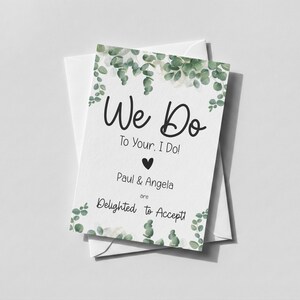 Puede incluir: Una tarjeta de respuesta de boda rectangular blanca con ilustraciones de hojas de eucalipto verdes en la parte superior e inferior. El texto dice: We Do To Your, I Do! Paul & Angela are Delighted to Accept! Un peque&ntilde;o coraz&oacute;n negro est&aacute; centrado.