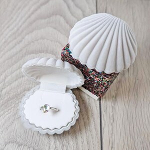 May include: A white velvet seashell ring box with a sparkly iridescent interior. The box is open and a silver ring with a clear iridescent stone is inside.