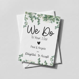 Puede incluir: Una tarjeta de respuesta de boda rectangular blanca con ilustraciones de hojas de eucalipto verdes en la parte superior e inferior. El texto dice: We Do To Your, I Do! Paul & Angela are Delighted to Accept! Un peque&ntilde;o coraz&oacute;n negro est&aacute; centrado.