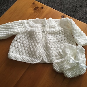 May include: A white knitted baby cardigan and matching booties with a white satin bow.
