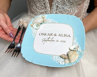 May include: A light blue ceramic plate with a white center decorated with seashells and a gold inscription that reads "OSKAR & ALISA September 16, 2026".