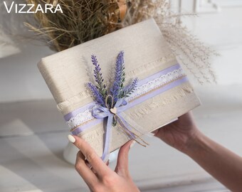 May include: A gift wrapped in beige linen fabric with a lavender flower bouquet, a purple ribbon, and a white lace ribbon. The gift is tied with a twine bow and a small wooden heart.