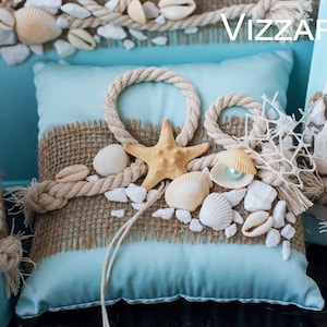 May include: A light blue pillow with a burlap and rope trim, decorated with seashells, a starfish, and white pebbles. The pillow is on a blue surface with the word "VIZZARA" in white text.
