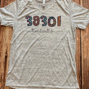 Op de afbeelding: Een grijs heather v-hals T-shirt met een kleurrijke afbeelding van de nummers 39301 en de tekst "Meridian, MS".