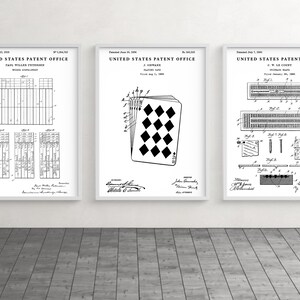 May include: Three framed patent prints. The prints are black and white, featuring detailed diagrams and text. The central print showcases a playing card design. The other two prints display technical drawings. The frames are white, and the background is a wooden floor.