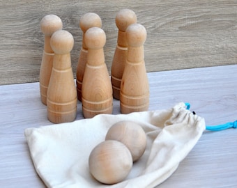 May include: A wooden skittles game set. Six wooden pins stand upright, with two wooden balls and a canvas bag. The pins are light brown with a rounded top and a wider base. The canvas bag is off-white, and the drawstring is light blue.