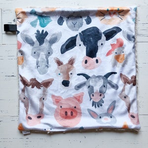 May include: A white baby blanket featuring watercolor illustrations of farm animals, including a cow, pig, and donkey. The blanket has a soft, plush border and a black strap. The overall design is a whimsical, colorful pattern.