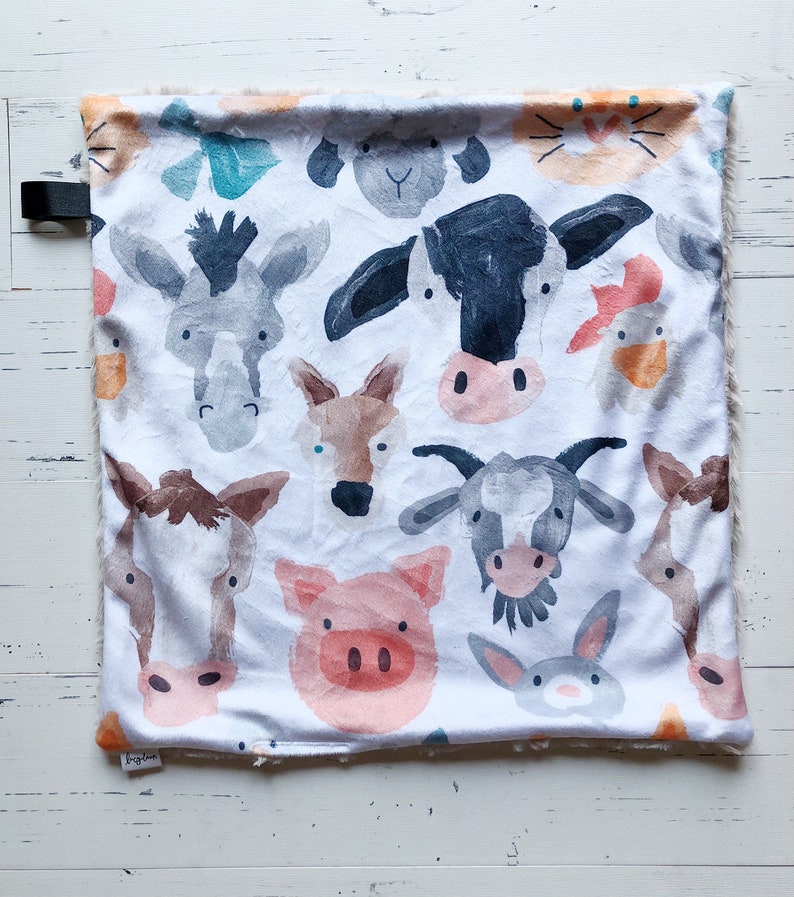 May include: A white baby blanket featuring watercolor illustrations of farm animals, including a cow, pig, and donkey. The blanket has a soft, plush border and a black strap. The overall design is a whimsical, colorful pattern.