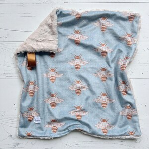 May include: A soft, blue baby blanket with a white faux fur border. The blanket features a repeating pattern of brown bees on a light blue background. The blanket has a small tag with the word "Beehive" on it.