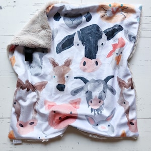 May include: A white baby blanket featuring watercolor illustrations of farm animals, including a cow, pig, and horse. The blanket has a soft, plush, tan border. The blanket is folded on a white wooden surface.