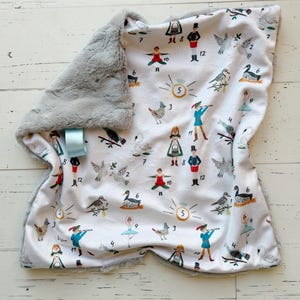 May include: A white baby blanket with a gray plush border. The blanket features a whimsical print of figures, birds, and numbers. A blue ribbon is attached to the blanket. The blanket is on a white wooden surface.