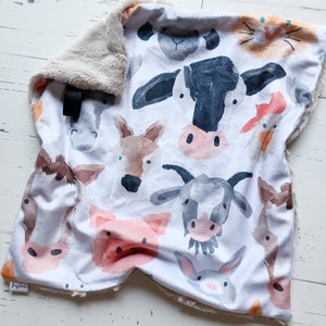 May include: A white baby blanket featuring watercolor illustrations of farm animals, including a cow, pig, and goat. The blanket has a soft, plush, tan border. A black tag is attached to the blanket.