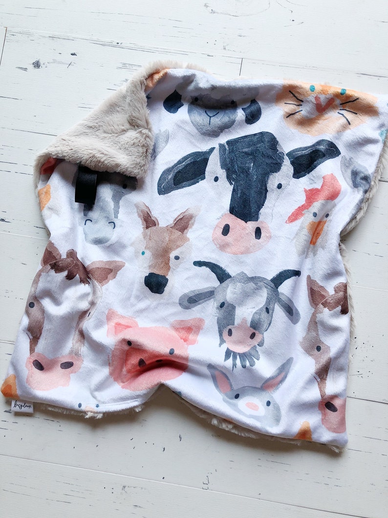 May include: A white baby blanket featuring watercolor illustrations of farm animals, including a cow, pig, and goat. The blanket has a soft, plush, tan border. A black tag is attached to the blanket.