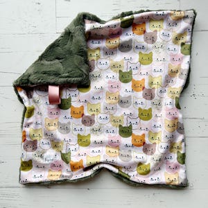 May include: A baby blanket featuring a pattern of cat faces in various colors on a white background. The blanket has a dark green, plush border and a pink satin ribbon. The blanket is made of soft material.