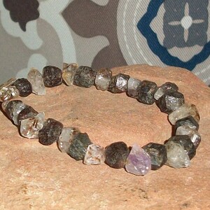 Könnte beinhalten: Ein Armband aus schwarzen und klaren Edelsteinen mit einem silbernen Verschluss. Das Armband besteht aus facettierten Steinen und liegt auf einem braunen Stein.