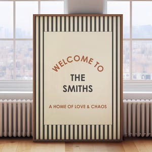 May include: Framed wall art with the text "WELCOME TO THE SMITHS" in a serif font. The artwork has a cream-colored background with a brown frame and black and brown striped border. The phrase "A HOME OF LOVE & CHAOS" is below the family name.