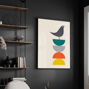 May include: A framed wall art print featuring a dark gray bird perched atop a vertical stack of four semi-circles in teal, gray, orange, and yellow, set against a cream background. The artwork is displayed in a black frame on a dark wall.