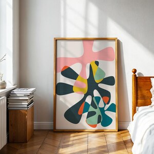 May include: A framed vertical wall art print featuring an abstract composition of overlapping organic shapes in shades of pink, dark charcoal, mustard yellow, teal, and orange against a light cream background, enclosed in a light wood frame.