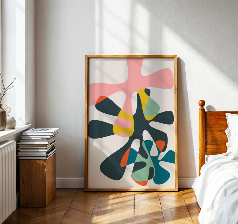 May include: A framed vertical wall art print featuring an abstract composition of overlapping organic shapes in shades of pink, dark charcoal, mustard yellow, teal, and orange against a light cream background, enclosed in a light wood frame.