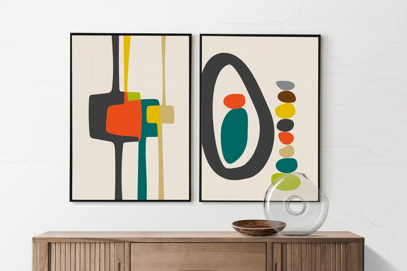 May include: Two framed wall art prints featuring abstract geometric shapes in black, orange, teal, yellow, and beige. The left print displays vertical lines and block forms, while the right print shows a large oval enclosing a teal shape and a stack of pebbles.