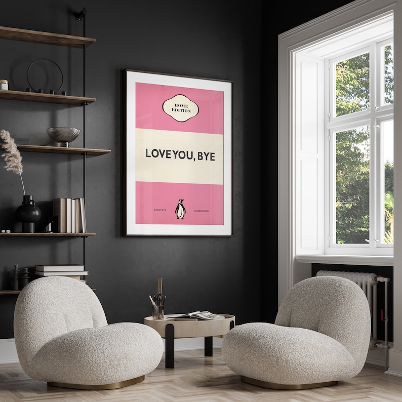 May include: A pink and cream book cover with the text "LOVE YOU, BYE" and a penguin illustration. The cover is framed and hanging on a black wall in a room with two white fluffy armchairs and a small wooden table.
