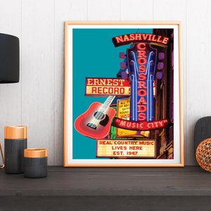 May include: A framed poster with a Nashville theme. The poster features neon signs for "Crossroads" and "Music City", a red guitar, and the text "Real Country Music Lives Here Est. 1947". The poster is on a dark surface with a lamp and candles.