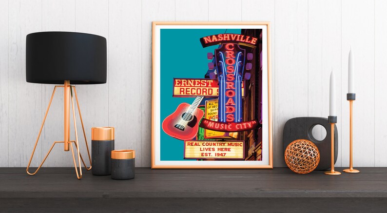 May include: A framed poster with a Nashville theme. The poster features neon signs for "Crossroads" and "Music City", a red guitar, and the text "Real Country Music Lives Here Est. 1947". The poster is on a dark surface with a lamp and candles.